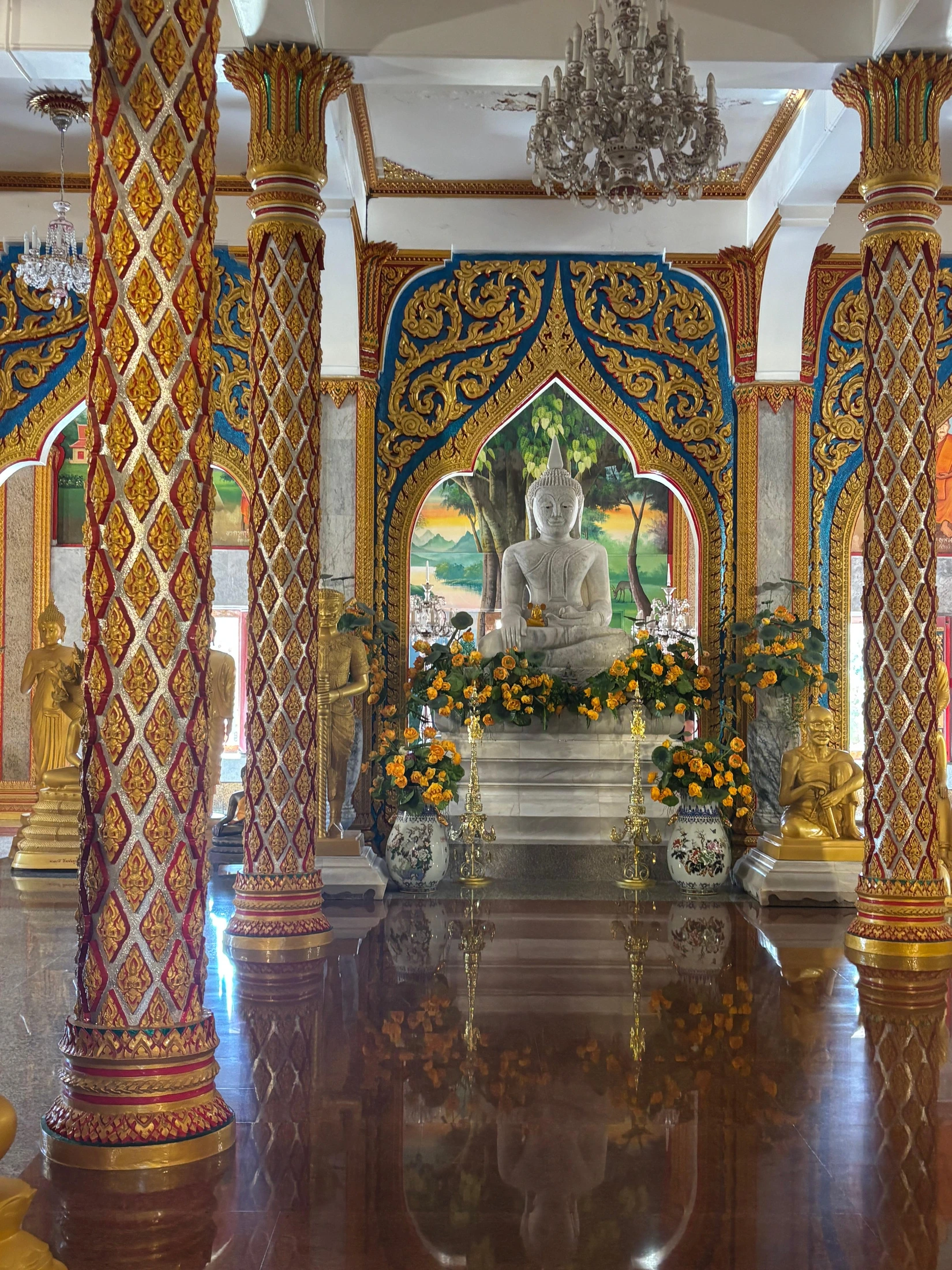 White Buddha statue among gilded columns, yellow flowers and blue arches inside a temple.