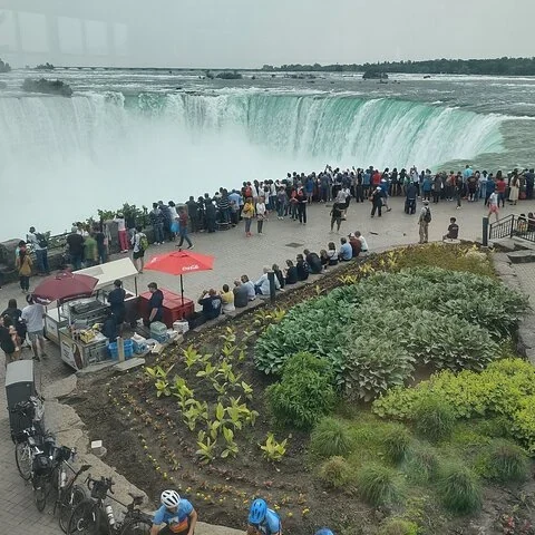 Des personnes se promènent et se rassemblent près d’une cascade incurvée ; jardins, bancs et stands avec parasols occupent le premier plan.