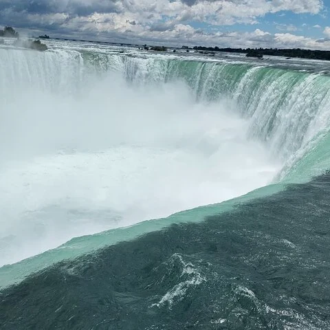 L’eau forme une courbe au bord d’une grande cascade et tombe dans une zone enveloppée de brume, sous un ciel nuageux.
