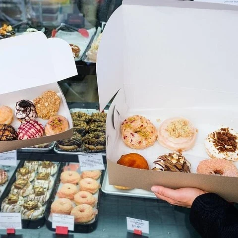Eine Person hält eine offene Schachtel mit Donuts vor einer Gebäckvitrine.