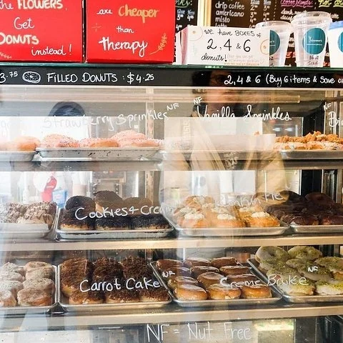 Eine Vitrine zeigt Donuts auf mehreren Ebenen, mit auf das Glas geschriebenen Namen und Schildern darüber.