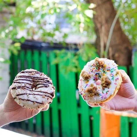 Zwei Hände halten Donuts im Freien: einer trägt dunkle Linien, der andere einen hellen Belag mit bunten Verzierungen.