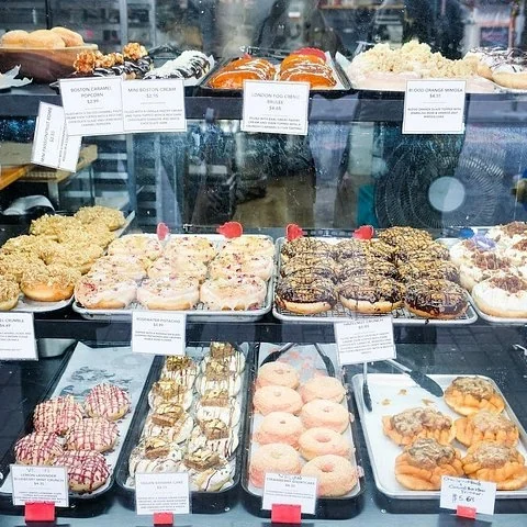 Eine Vitrine enthält auf mehreren Ebenen Bleche mit unterschiedlich belegten Donuts und dazugehörigen Etiketten.