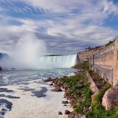 The falls and their mist beside a rocky shore and buildings.