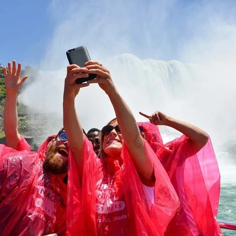 A group in pink ponchos poses in the mist while one person holds a phone overhead; a waterfall is visible behind them.