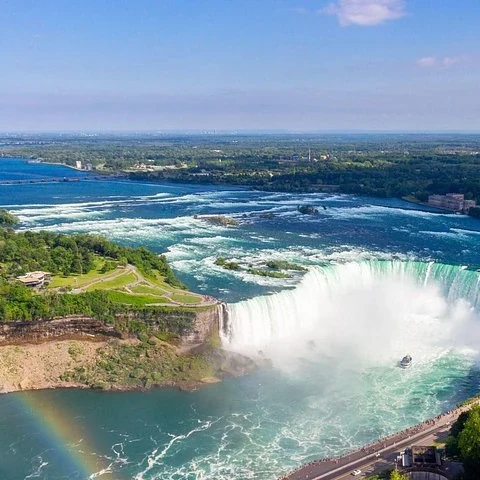 An elevated view shows a curved waterfall, a broad river, a small boat in the mist and a rainbow near the shore.