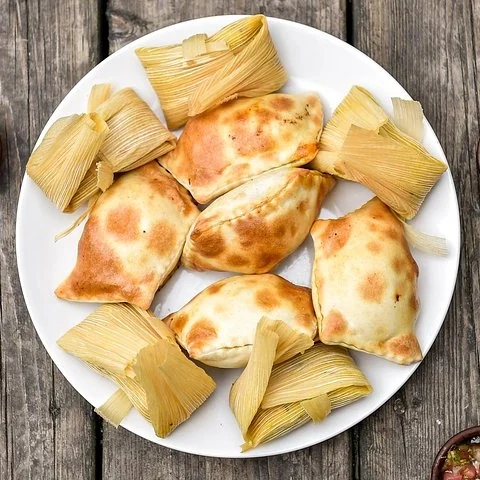 A round plate holds golden baked pastries and leaf-wrapped parcels on a wooden table.