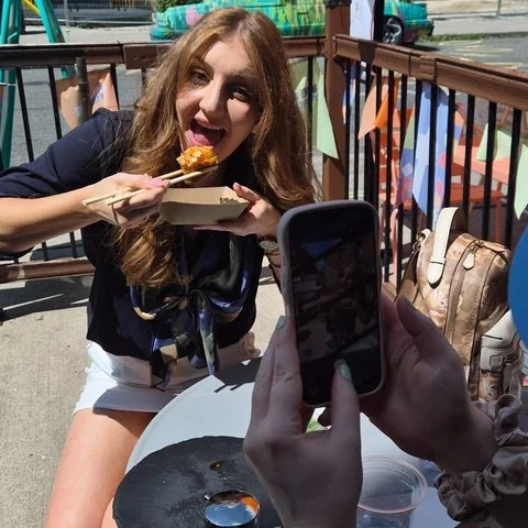 A woman brings a bite towards her mouth with chopsticks while another person points a phone camera at her.