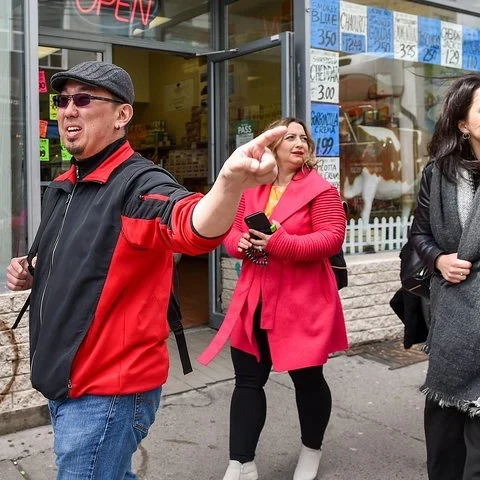 A man in a cap and a red-and-black jacket points to one side; two women stand beside him outside a shop.
