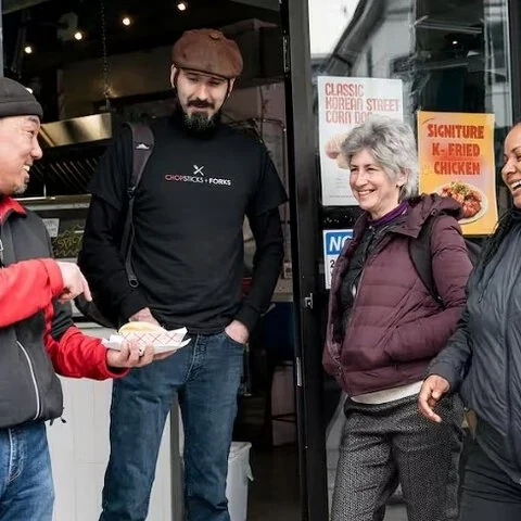 Three people talk outside a shop entrance, one holding a plate.