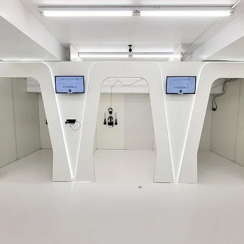 Mehrere weiße Kabinen mit Trennwänden; Bildschirme zeigen „STATION 2“ und „STATION 3“, in der Mitte hängen ein Headset und Controller.