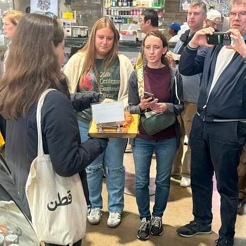 Eine Gruppe steht in einem Geschäft vor einer Person mit einem Tablett; eine weitere Person fotografiert.