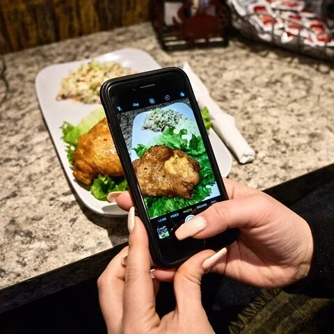 Hände halten ein Telefon, um einen Teller mit grünen Blättern und einem goldbraunen Stück Essen zu fotografieren.