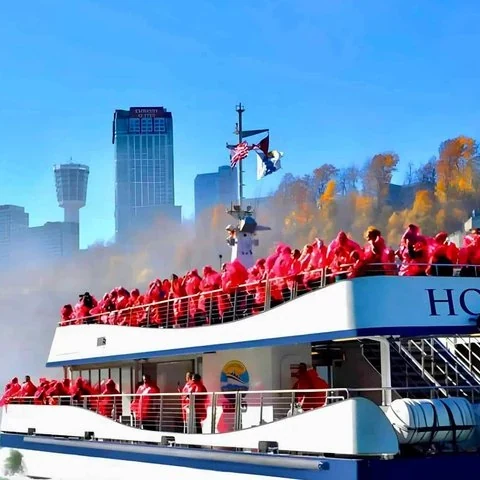 Un barco blanco lleva pasajeros con ponchos rojos en sus cubiertas; detrás se ven torres, edificios y árboles.