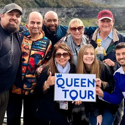 Un grupo posa al aire libre; dos personas sostienen un cartel blanco con las palabras QUEEN TOUR en azul.