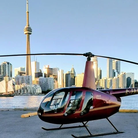 Ein weinroter Hubschrauber steht am Wasser; dahinter sind der CN Tower und hohe Gebäude zu sehen.