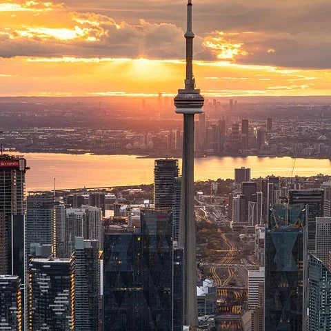 Der CN Tower ragt zwischen Gebäuden unter einem orangefarbenen Himmel auf; Sonnenlicht spiegelt sich im Wasser.