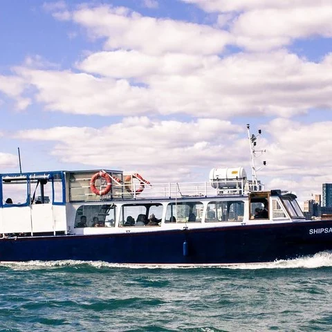 Ein Boot mit dunklem Rumpf und weißer Kabine fährt über das Wasser; mehrere Personen sind hinter den Fenstern zu sehen.