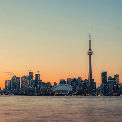 Die Skyline, eine Kuppel und ein hoher Turm heben sich vor einem orangefarbenen Himmel ab; im Vordergrund liegt Wasser.