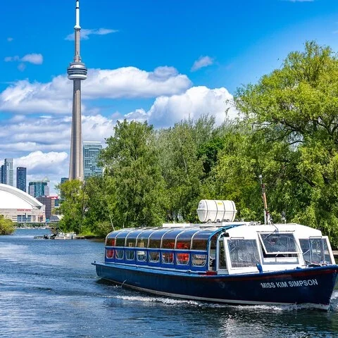 Ein blau-weißes Boot fährt an Bäumen vorbei, mit dem CN Tower im Hintergrund.