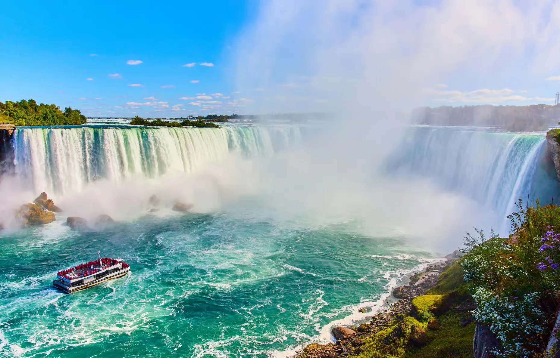 A boat sails in front of the falls, mist and a rainbow.